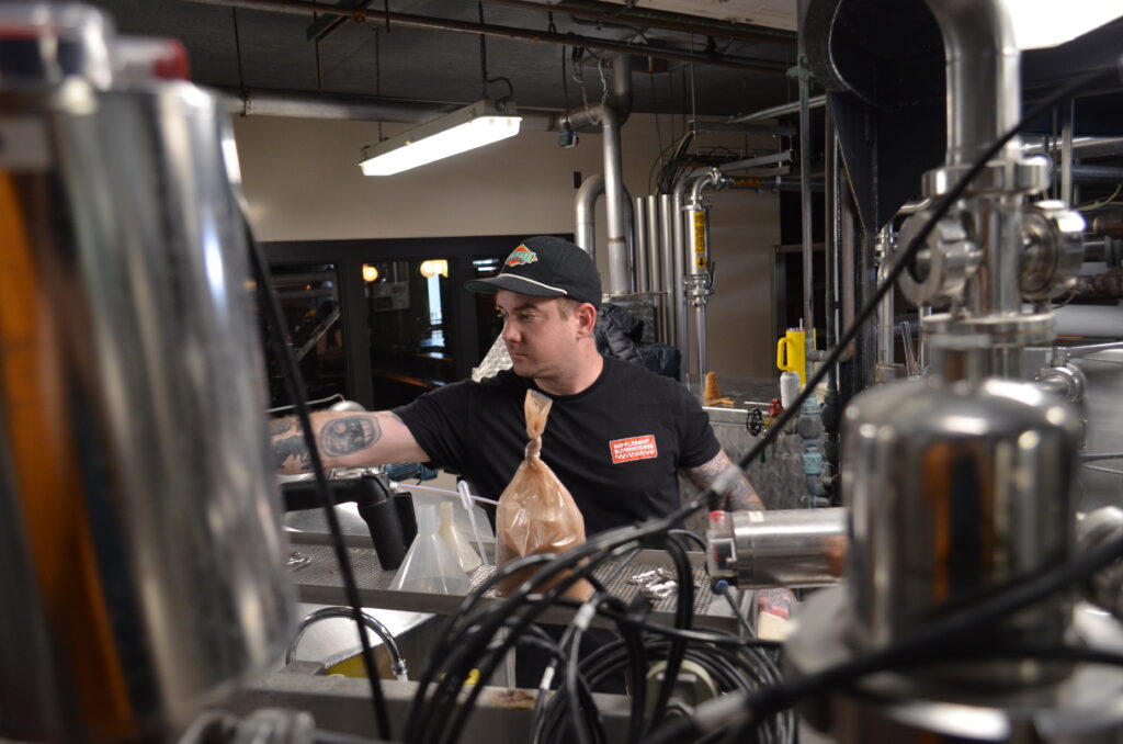 A person in a black cap and t-shirt works amidst industrial brewing equipment. They are adjusting machinery, surrounded by metal pipes and containers. A beige sack and coiled wires are visible in the foreground.