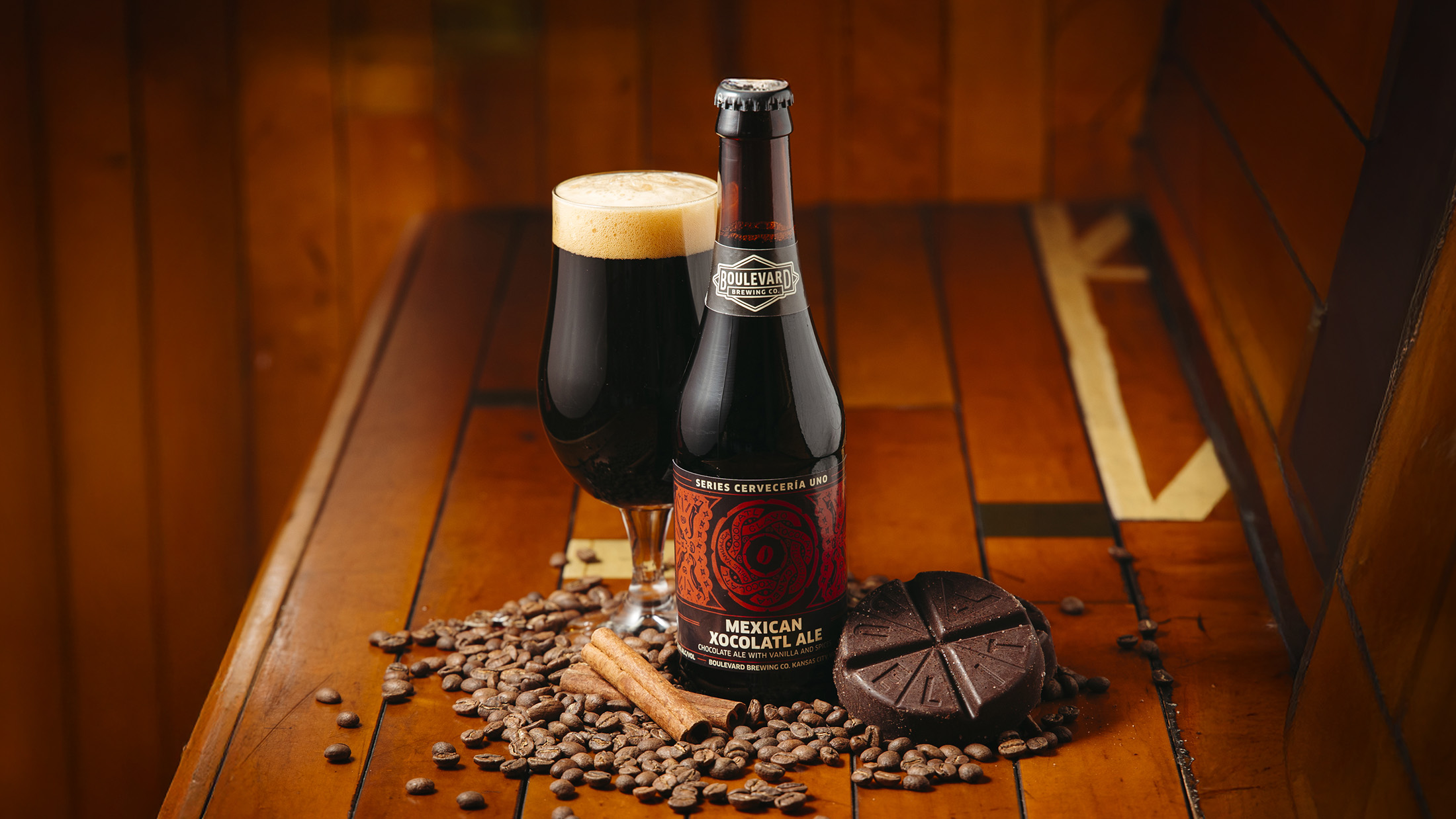 A bottle and glass of Mexican Xocolatl Ale sit on a wooden surface, surrounded by cocoa beans, chocolate pieces, and cinnamon sticks, creating a warm and inviting atmosphere.