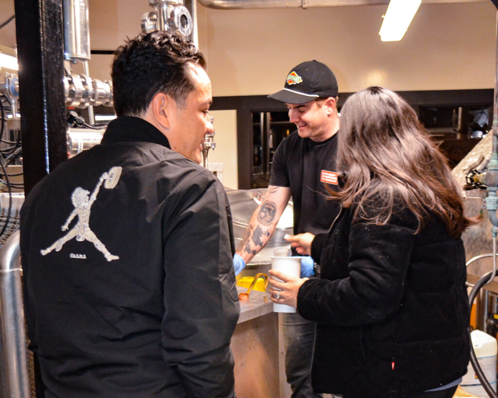 Three people are gathered in a workshop setting. One man, wearing a black cap and displaying tattoos, is engaging with the others, who are wearing black jackets. They seem to be involved in a collaborative activity.