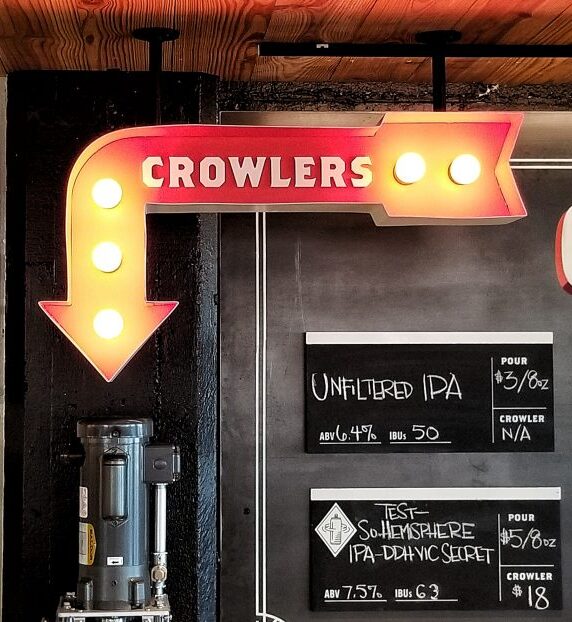 A pub's interior with a large red arrow sign labeled "CROWLERS" pointing downwards. Chalkboard menu shows various beers on tap. A poster on the left advertises fresh beer, and a row of beer cans is displayed below it.