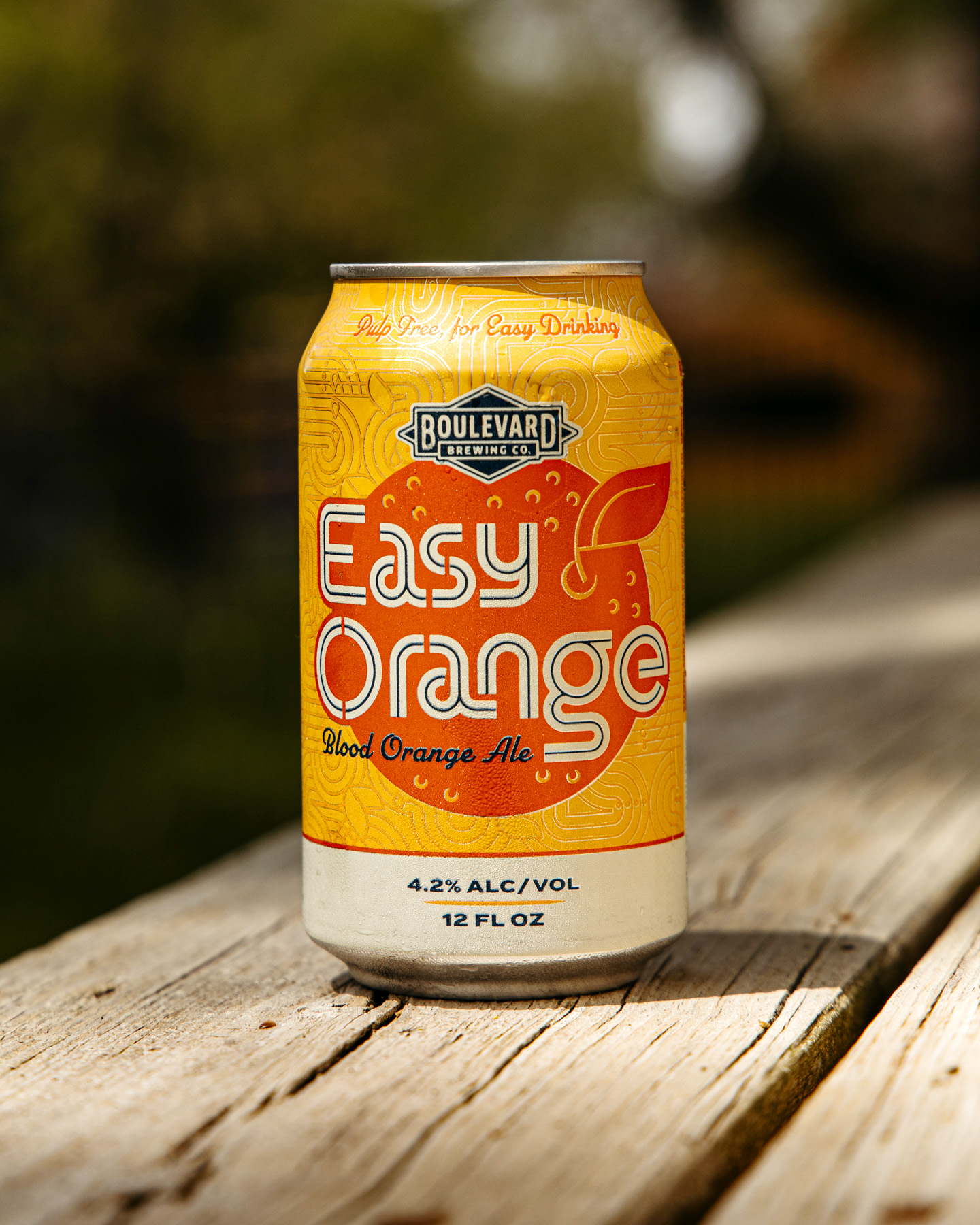 A can of Boulevard Brewing Co.’s Easy Orange Blood Orange Ale sits on a wooden surface outdoors. The can is bright orange with bold lettering and decorative citrus graphics.