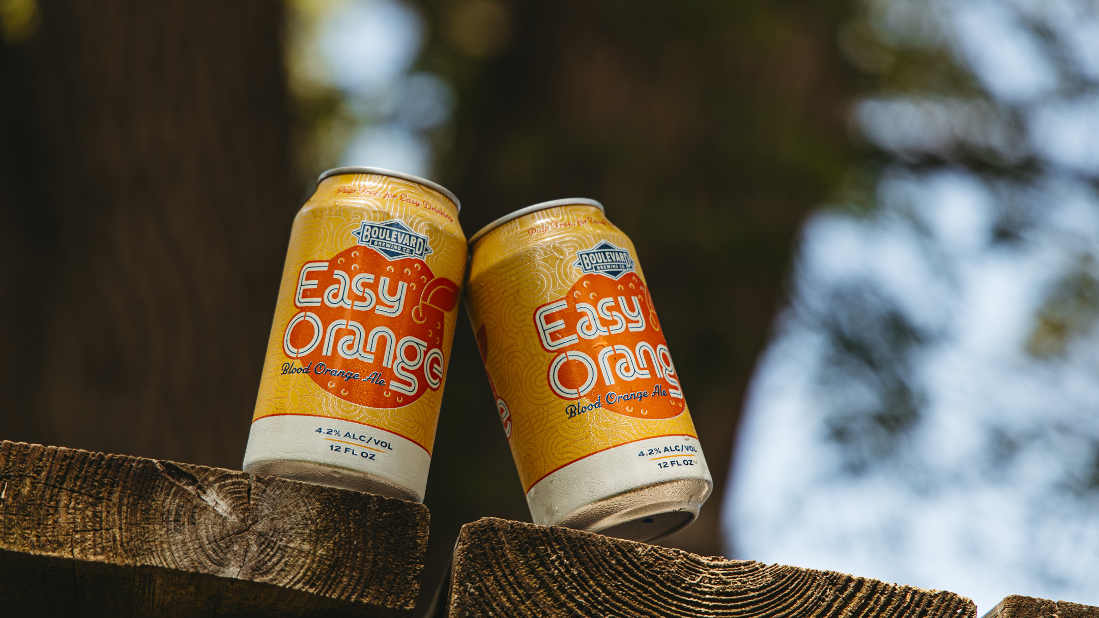 Two cans of Boulevard Brewing Co. Easy Orange beer rest on a wooden surface outdoors, with trees and sunlight blurred in the background. The cans are orange and yellow with blue accents.