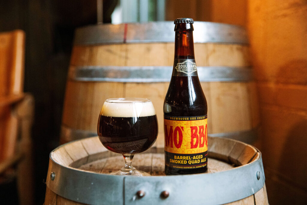 A bottle of MoxBBL barrel-aged smoked quad ale and a filled glass sit on a wooden barrel, with another barrel and warm amber lighting in the background.