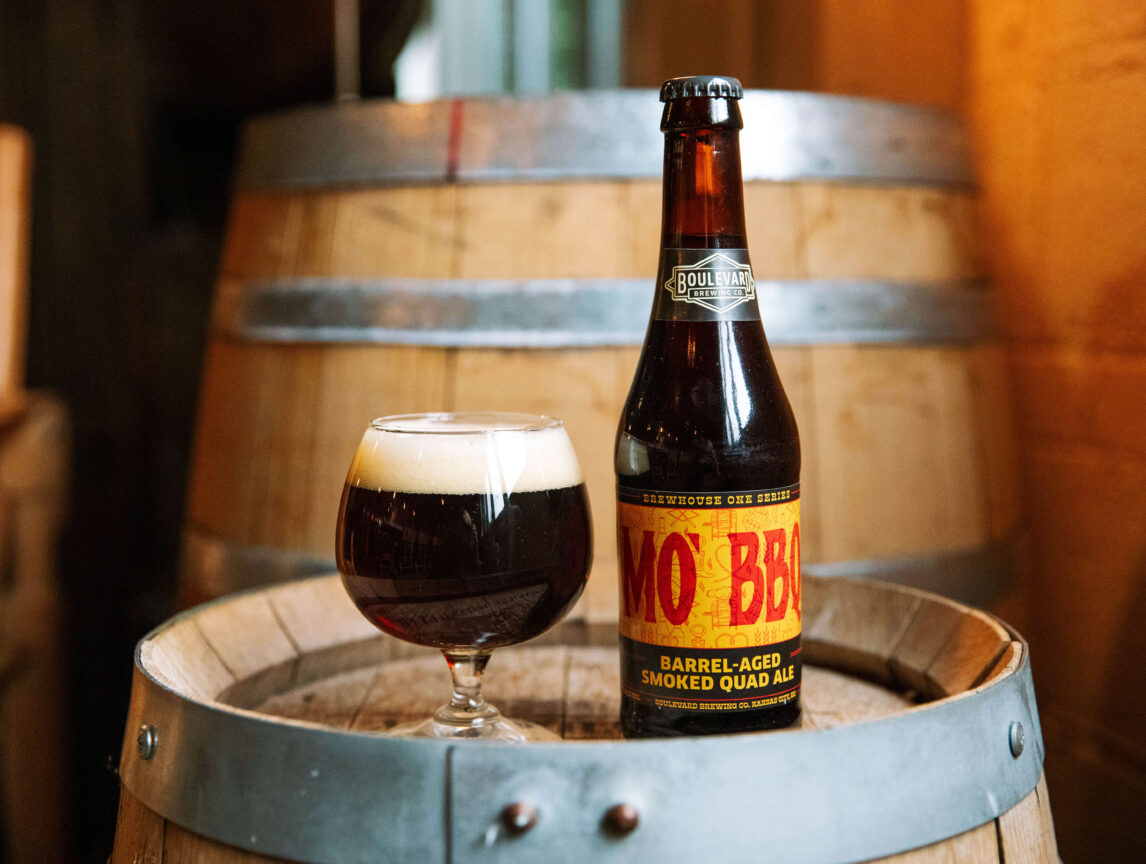 A bottle of MoxBBL barrel-aged smoked quad ale and a filled glass sit on a wooden barrel, with another barrel and warm amber lighting in the background.