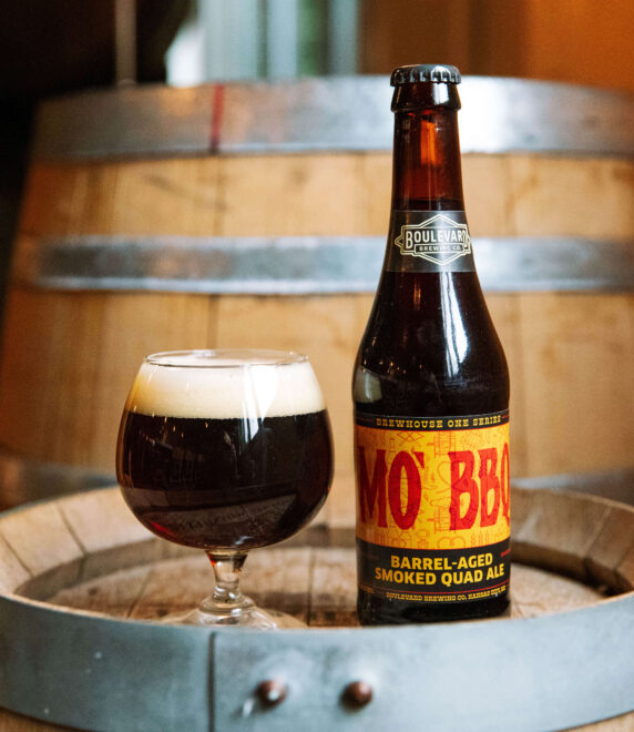 A bottle of MoxBBL barrel-aged smoked quad ale and a filled glass sit on a wooden barrel, with another barrel and warm amber lighting in the background.