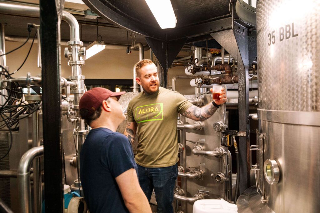 Two men stand in a brewery surrounded by metal tanks and pipes. One man, wearing a green "ALORA" shirt, holds up a glass of beer, while the other, in a cap and glasses, looks on.