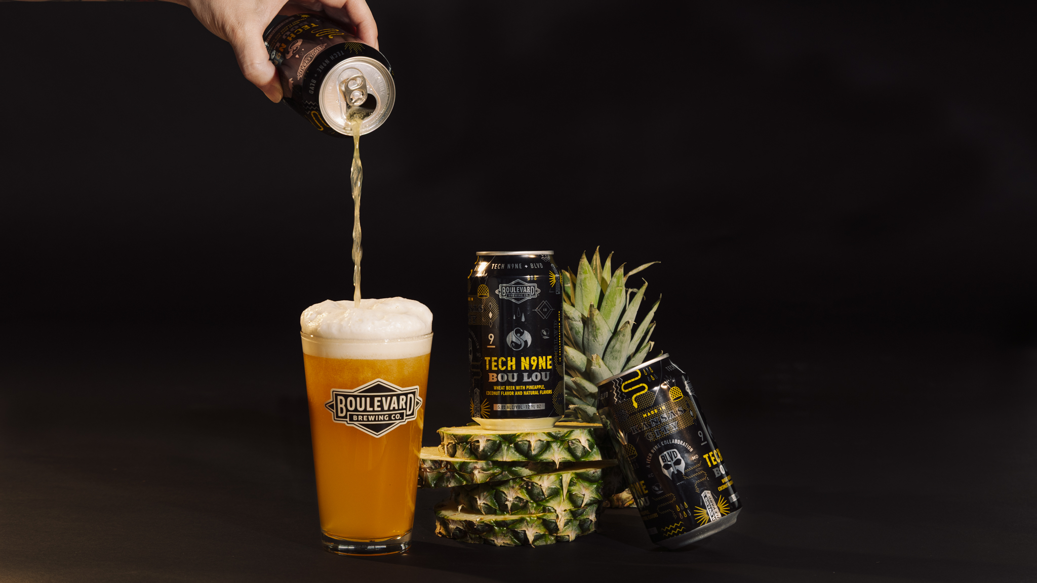 A hand pours beer from a can into a foamy Boulevard glass. Three cans, pineapple slices, and a pineapple crown are arranged on a dark background.