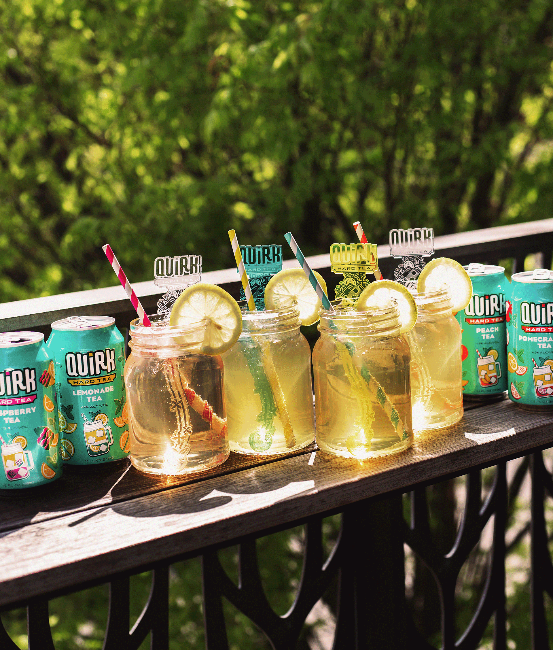 Four mason jars filled with refreshing lemonade IPA, each garnished with a lemon slice and colorful straw, are lined up on a sunlit railing. Behind them are six turquoise cans of Quirk Lemonade IPA. Lush greenery is visible in the background.