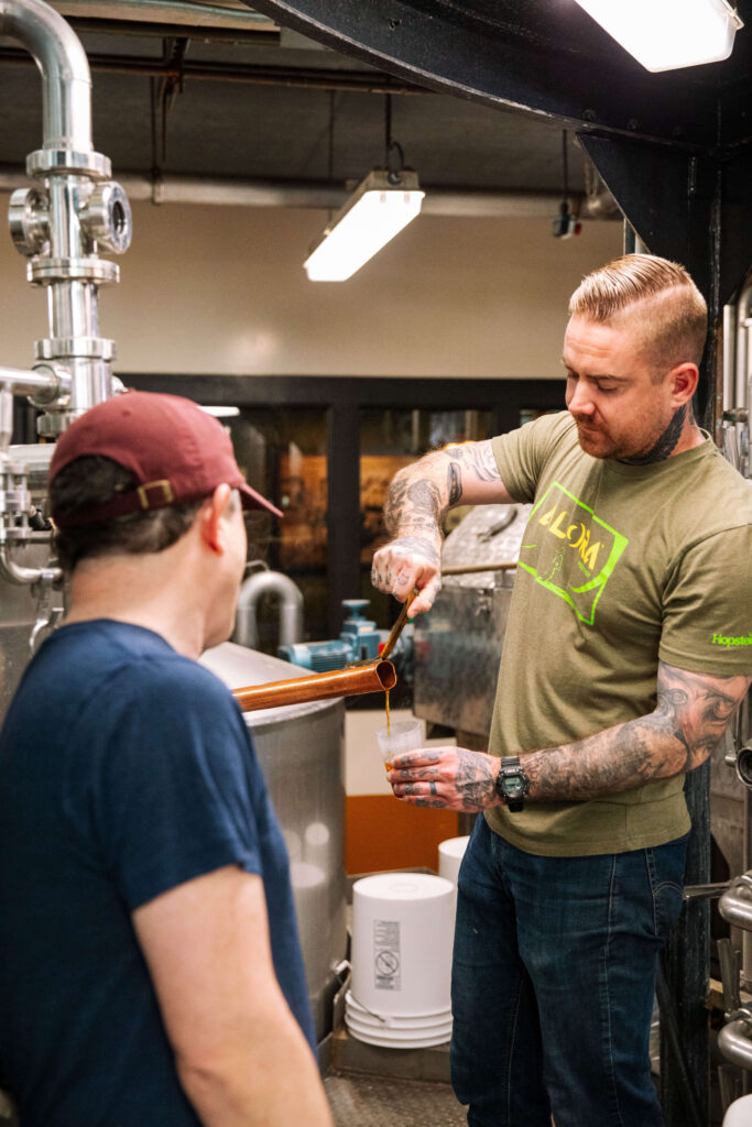 A man with tattoos pours a liquid into a glass while talking to another man in a brewery or industrial setting with pipes and tanks in the background.