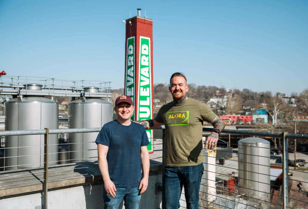 Two men stand smiling on an outdoor terrace with large brewing tanks and a tall red and green "Boulevard" sign behind them on a sunny day. One man holds a drink. Buildings and trees are visible in the background.