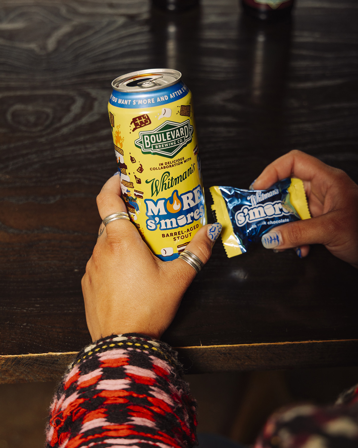 A person in a colorful sweater holds a can of Boulevard Brewing Co. More S’more beer in one hand and a Whitman’s s’more chocolate treat in the other, both on a dark wooden table.