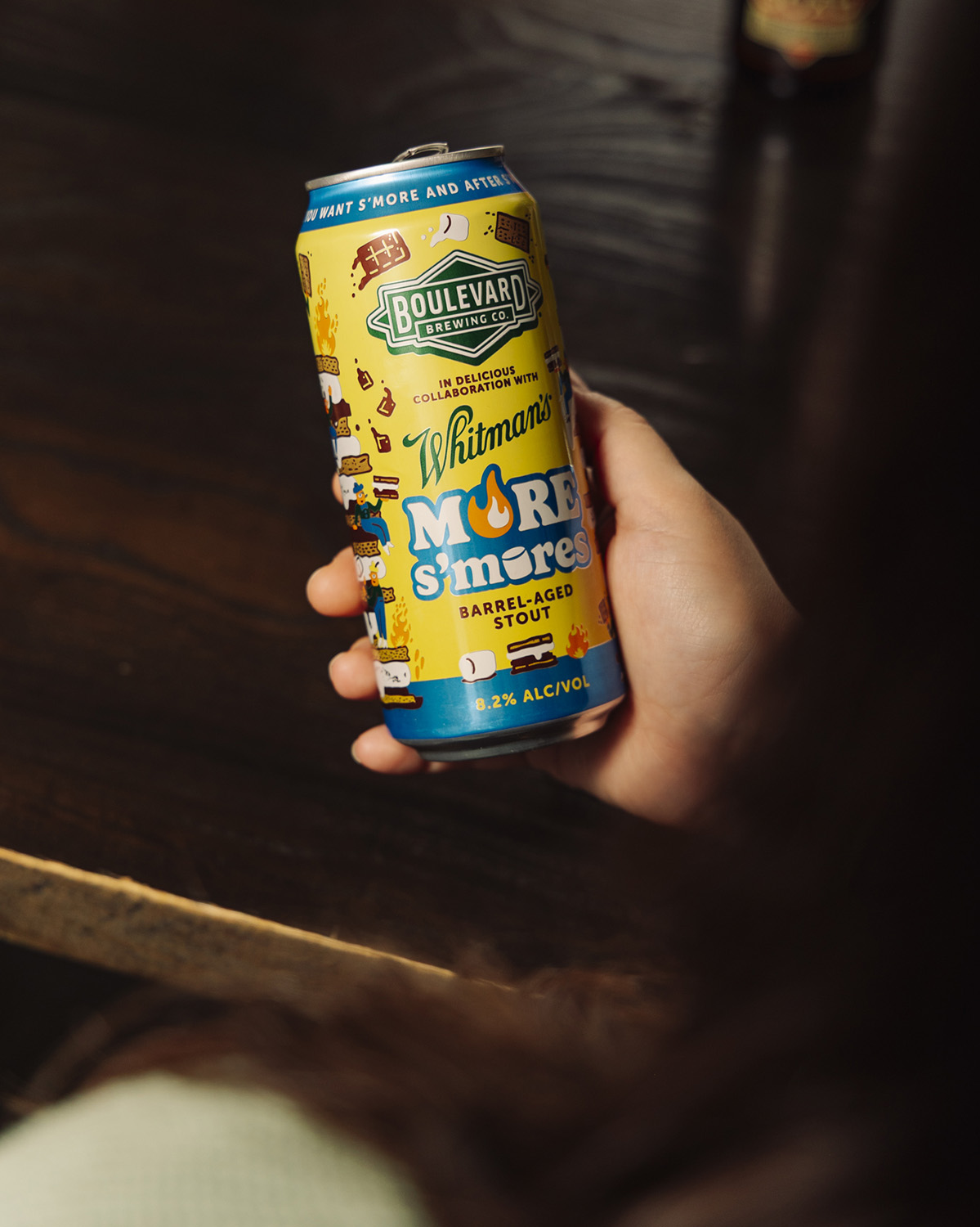 A person holds a can of Boulevard Brewing Co.'s More S'mores Barrel-Aged Stout, its colorful campfire and s'mores-themed artwork popping against a dark wooden table.