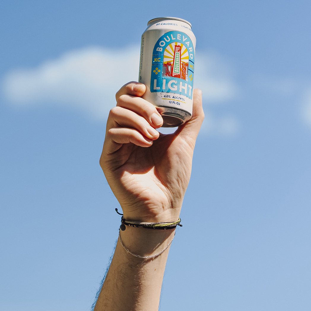 A hand wearing two bracelets holds up a can of Boulevard Brewing Co. Light beer against a bright blue Kansas City, Missouri sky with a small white cloud.