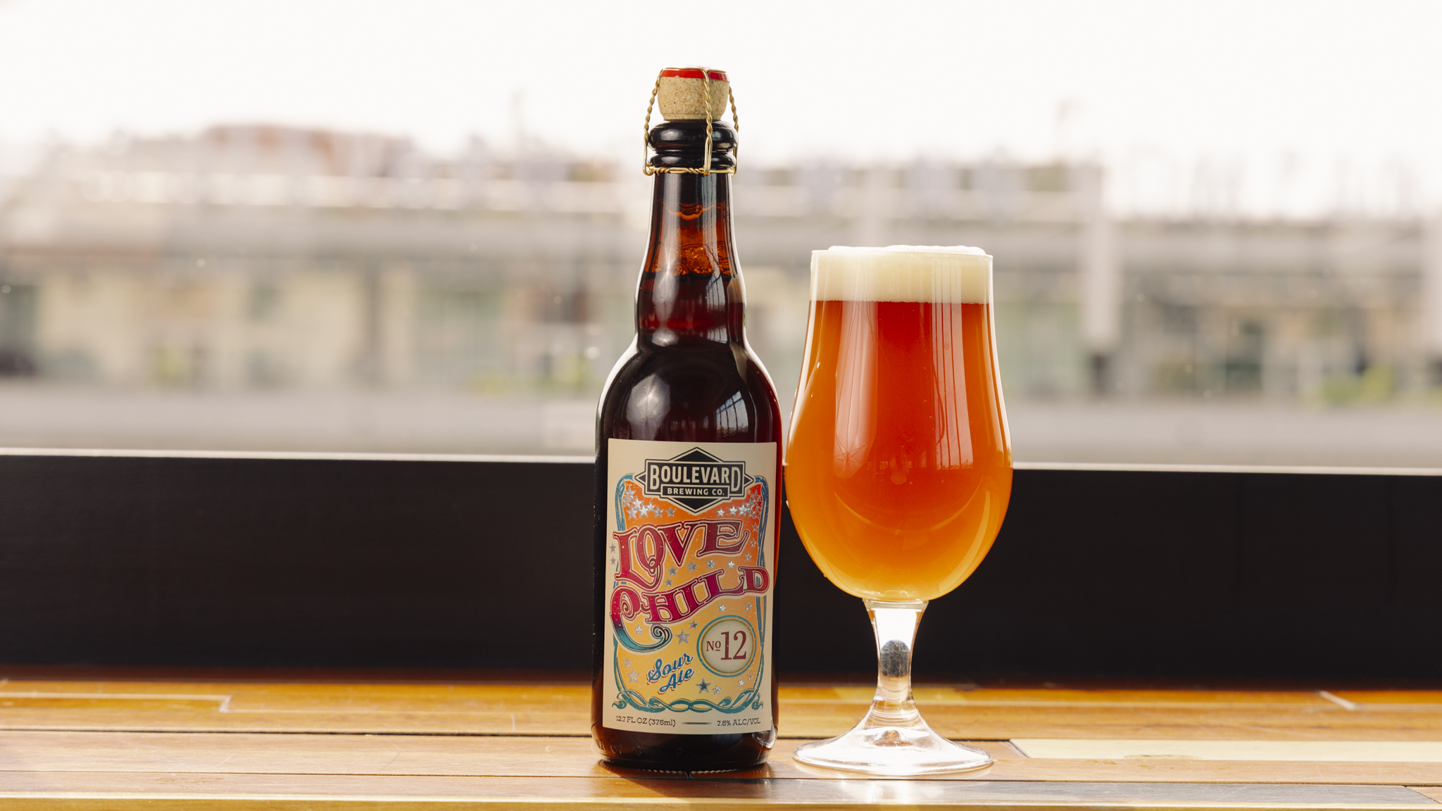 A bottle of Boulevard Love Child No. 12 beer stands next to a filled beer glass with a foamy head, set on a wooden surface with an urban background visible through a window.