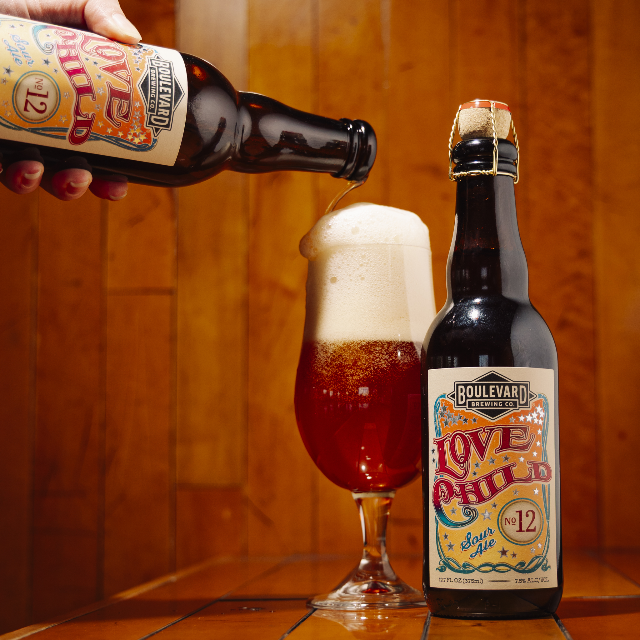 A hand pours Boulevard Brewing Co. Love Child No. 12 Sour Ale into a glass with a large foamy head. Another bottle of the same beer stands beside the glass against a warm, textured background.