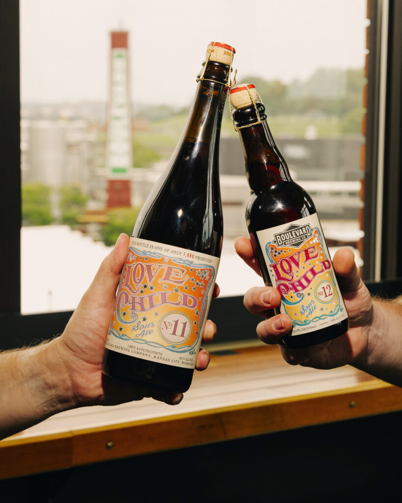Two hands clink bottles of Boulevard Brewing Co. Love Child No. 12 sour ale in front of a window, with a blurred view of industrial buildings and greenery outside.