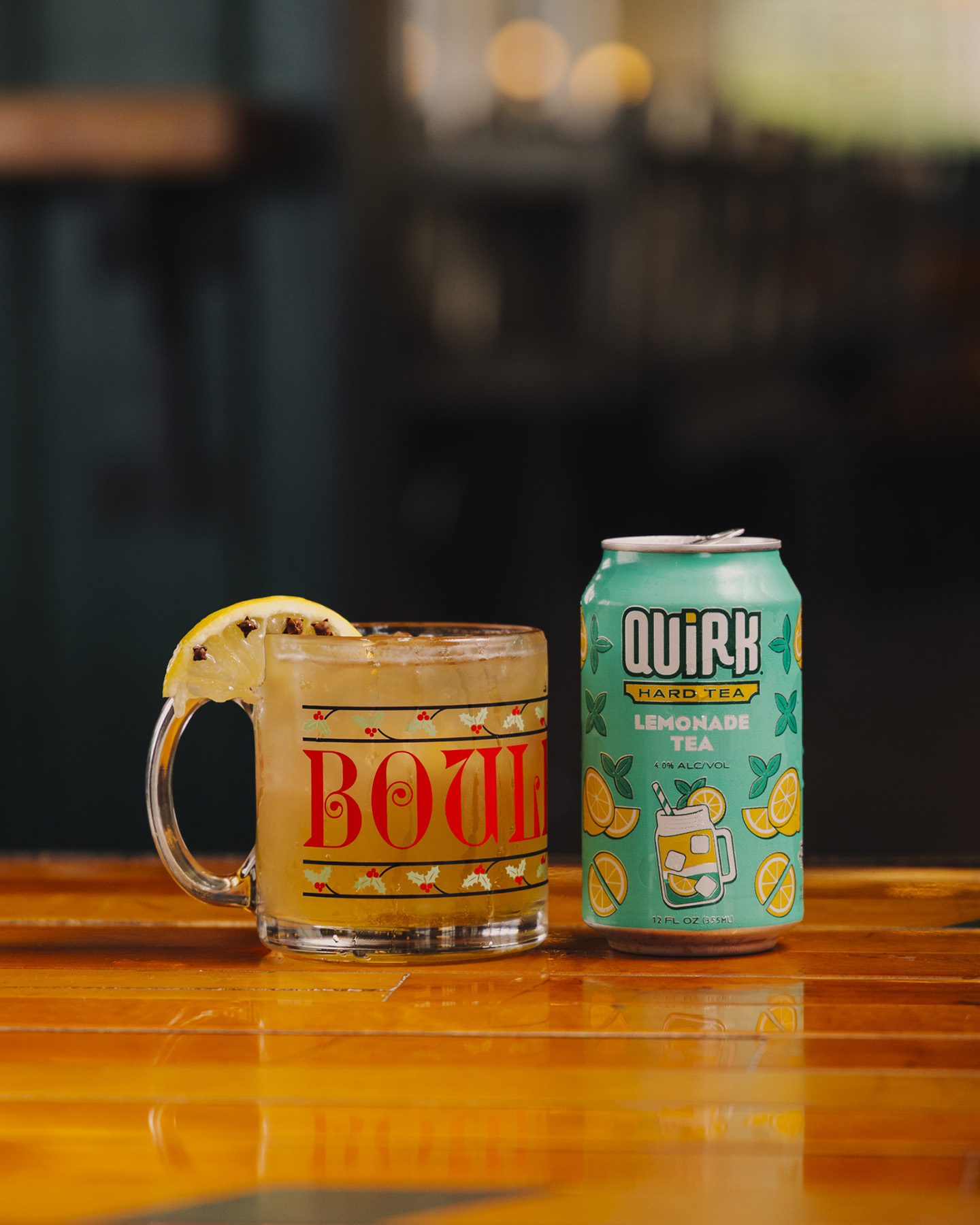 A mug with a lemon slice and a cold drink sits beside a teal can labeled “QUIRK Lemonade Tea” on a wooden table, with a blurred background.