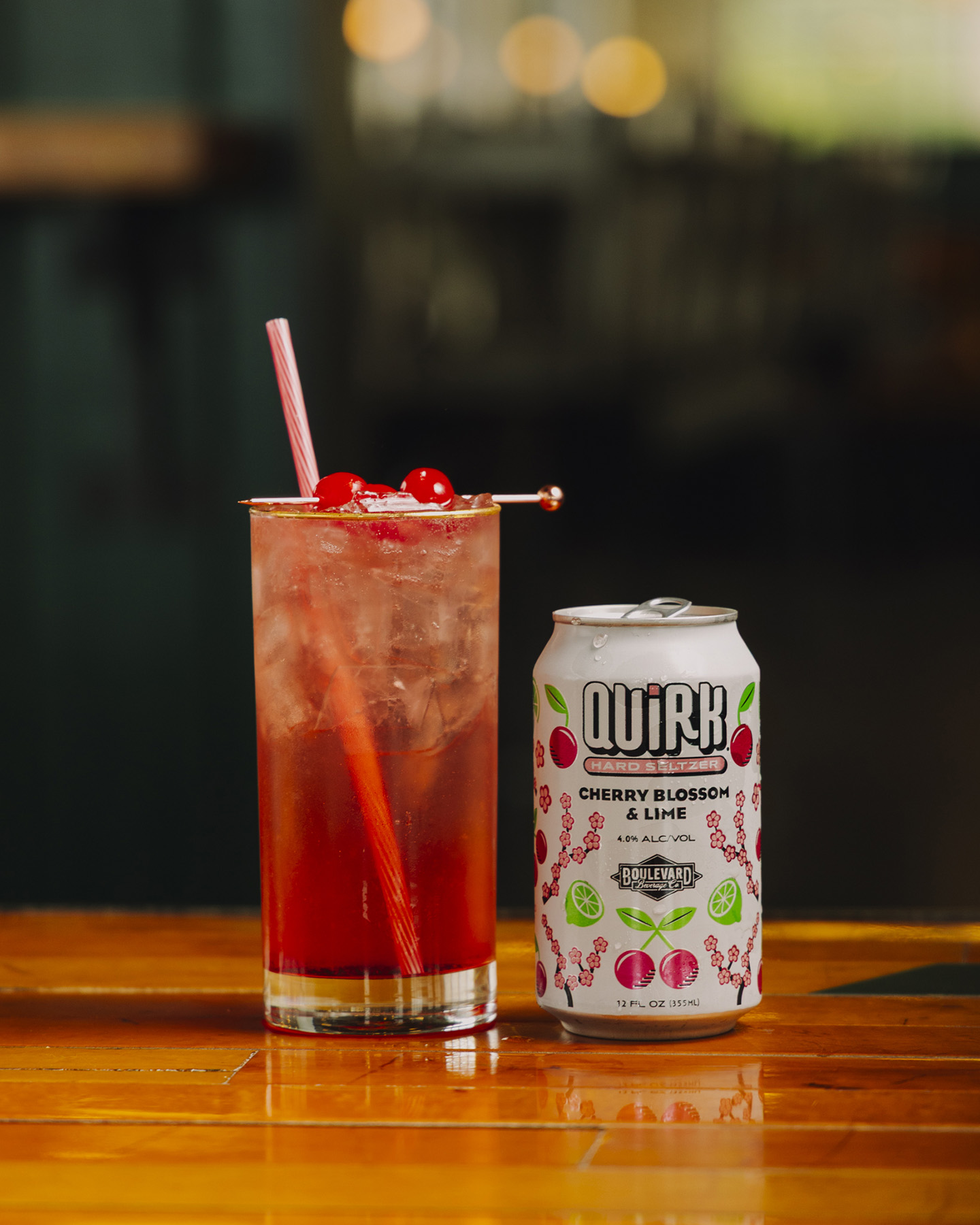 A tall glass of pink cherry blossom and lime drink with ice, garnished with cherries and a striped straw, sits next to a matching can labeled "Quirk Cherry Blossom & Lime" on a wooden table.