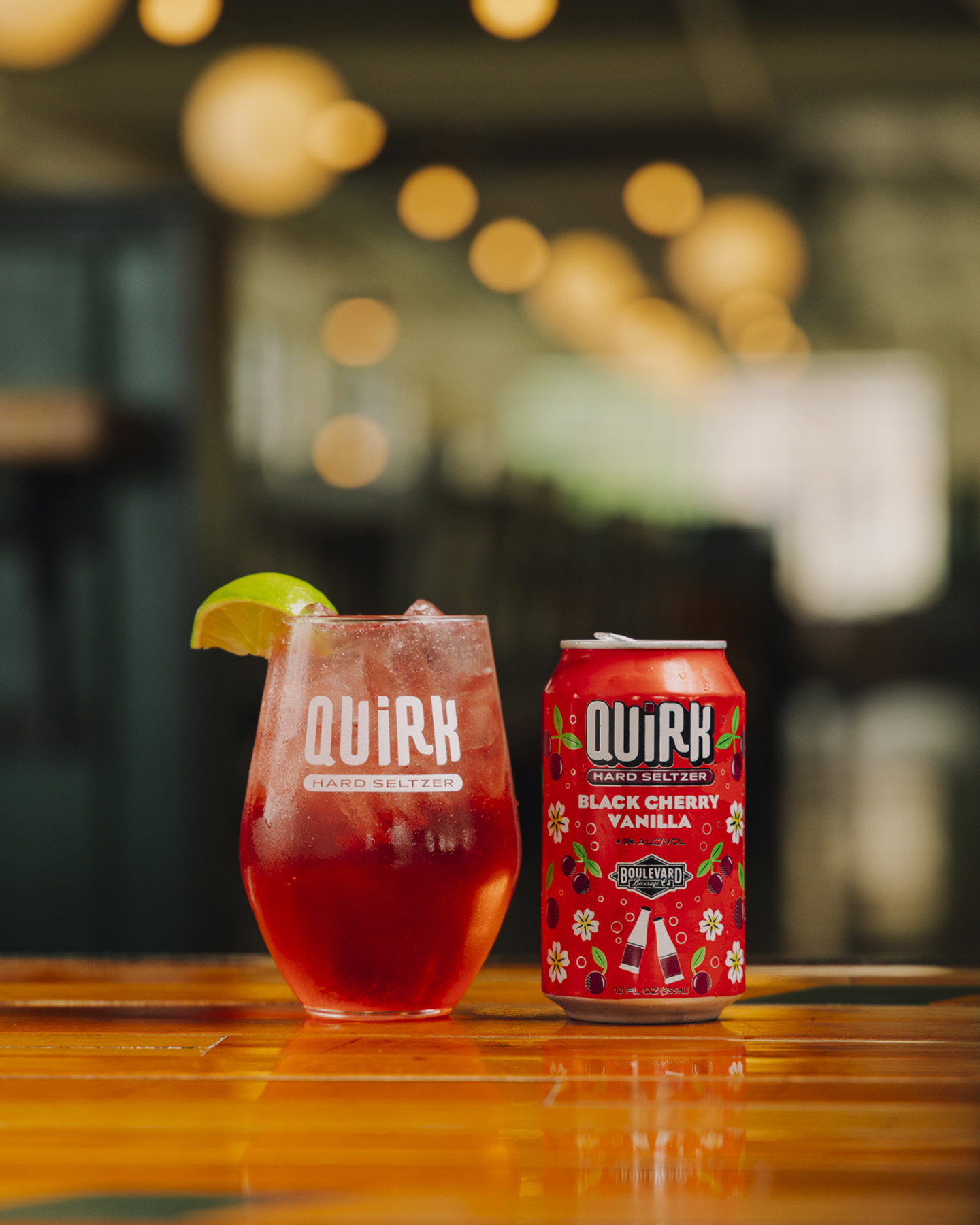 A glass of red sparkling drink with ice and a lime wedge sits next to a colorful can labeled "Quirk Hard Seltzer Black Cherry Vanilla" on a wooden surface, with blurred lights in the background.