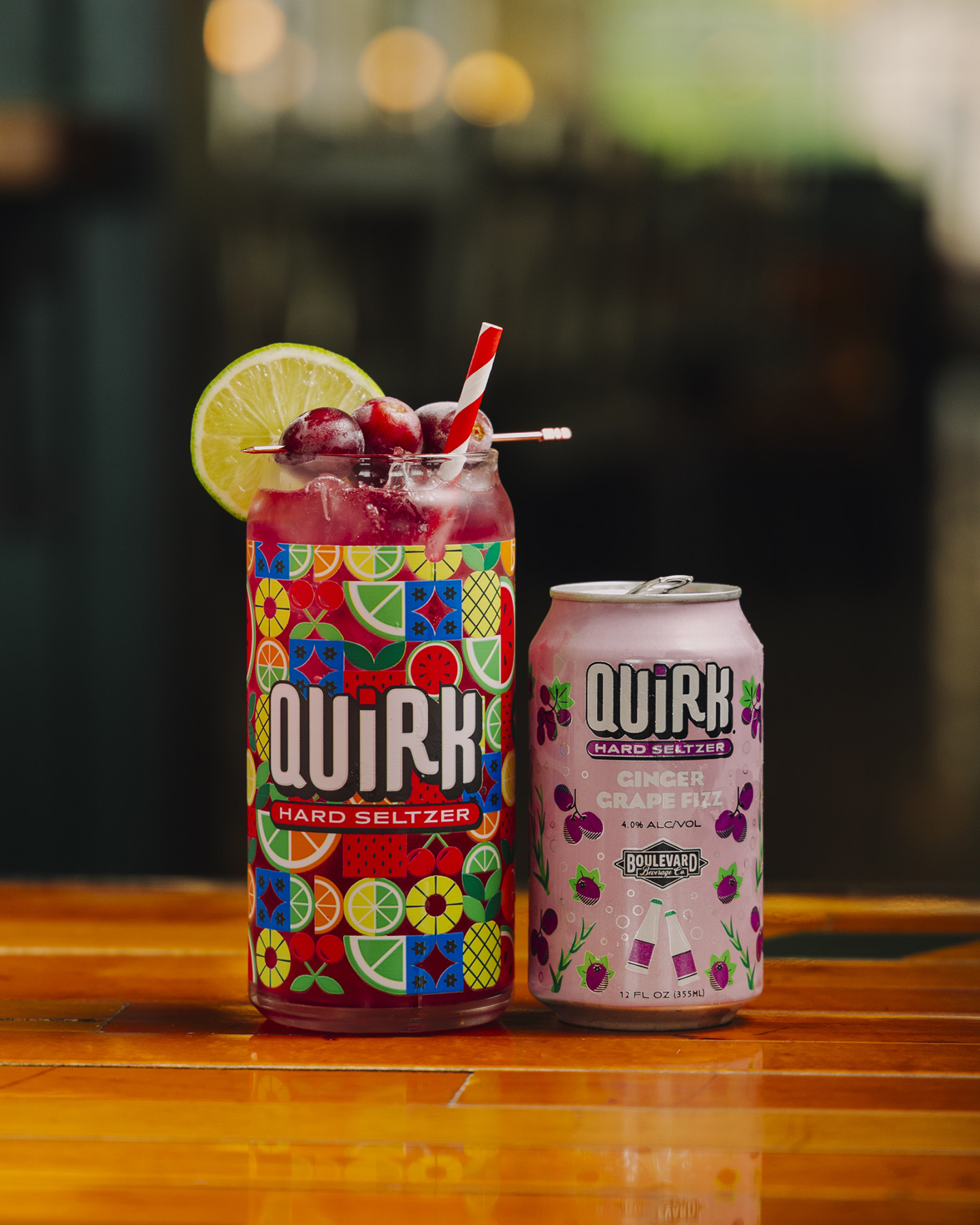 A glass decorated with colorful fruit patterns filled with a pink drink, ice, lime slices, and cherries sits next to a can of Quirk Hard Seltzer Cranberry Grape Fizz by Boulevard Brewing Co. on a wooden surface.