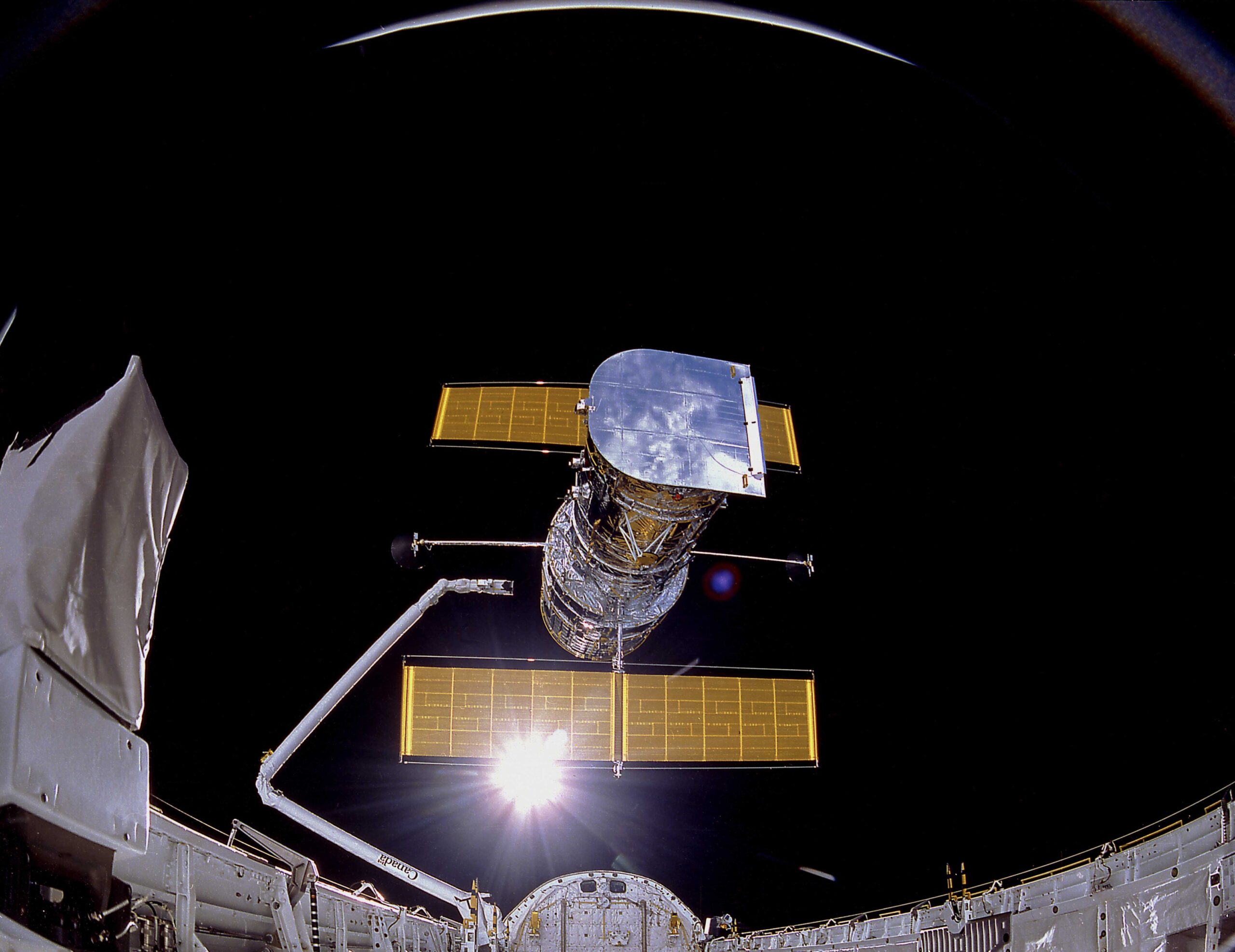 The Hubble Space Telescope is seen suspended above a space shuttle, with its solar panels extended, against the blackness of space and the bright glow of the sun in the background.
