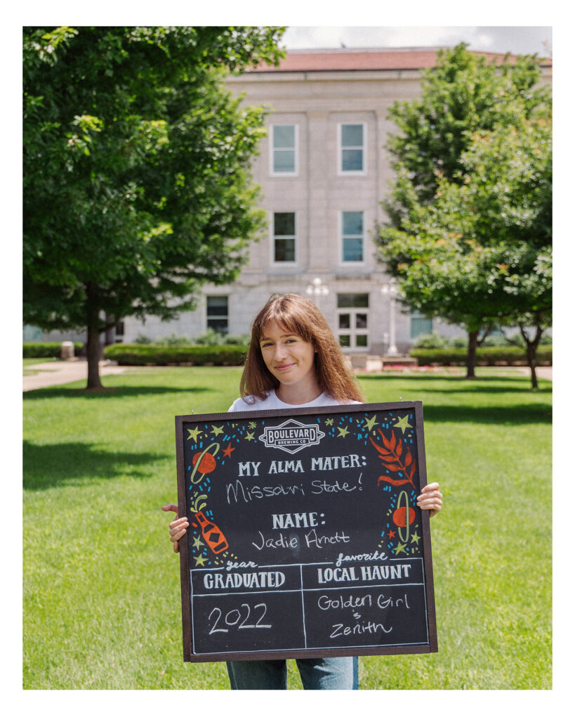A woman stands on grass in front of a campus building, holding a decorated chalkboard sign that reads: “Missouri State! Name: Jackie Merritt. Graduated: 2022. Favorite Local Haunt: Golden Girl, Zenith.” Trees are in the background.