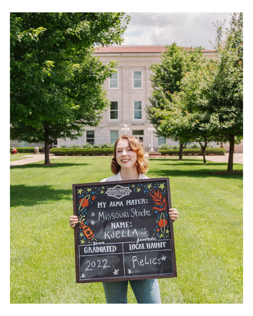 A person with shoulder-length hair stands smiling on a sunny lawn, holding a decorated chalkboard sign with graduation details in front of a large academic building.