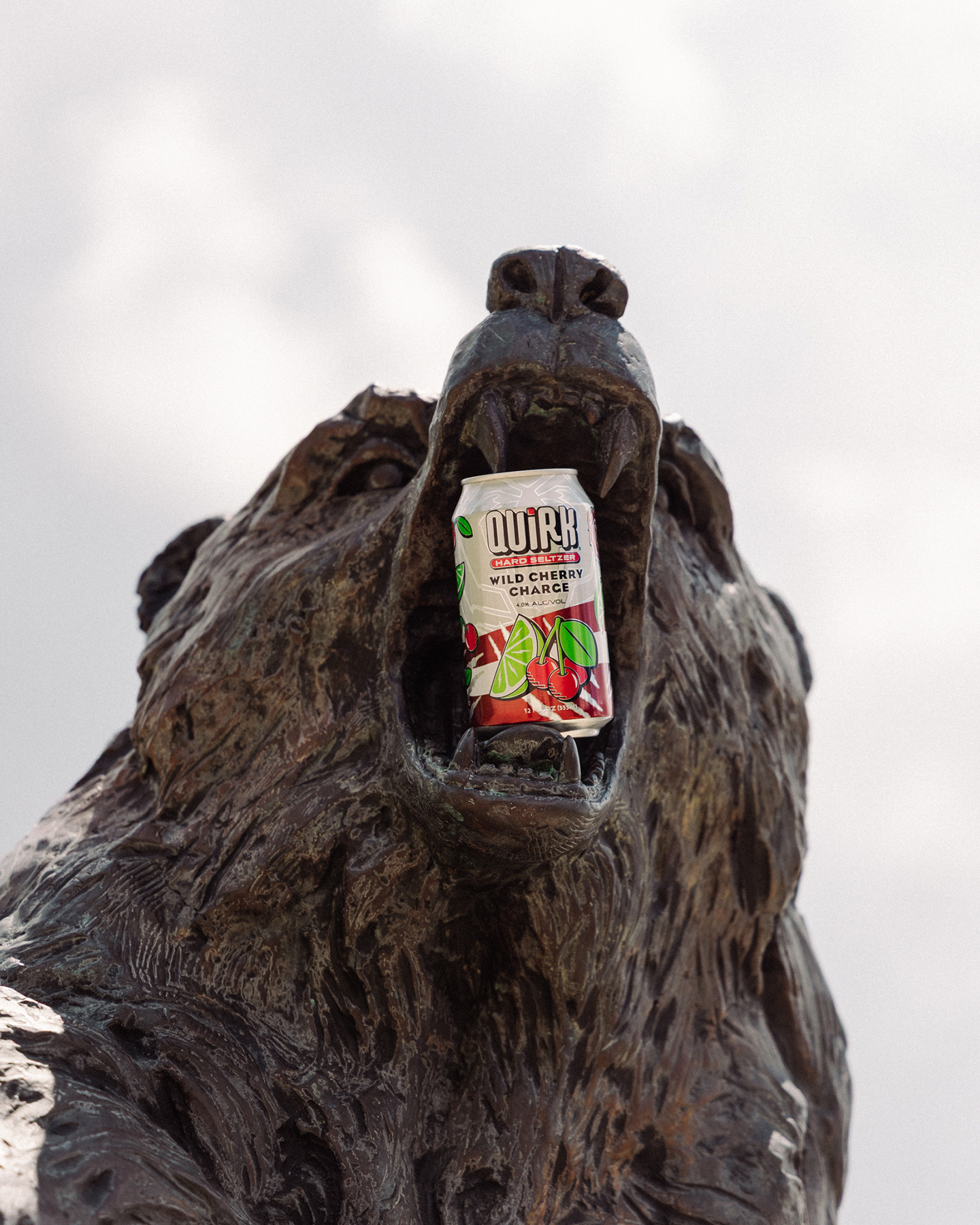 A close-up of a bear statue with its mouth open, holding a can of Quirk Hard Seltzer Wild Cherry Charge between its teeth against a cloudy sky background.