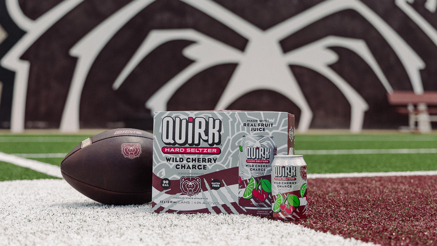 A box and can of Quirk Hard Seltzer Wild Cherry Charge sit on a football field next to a brown football, with a bear mascot graphic and bench in the background.