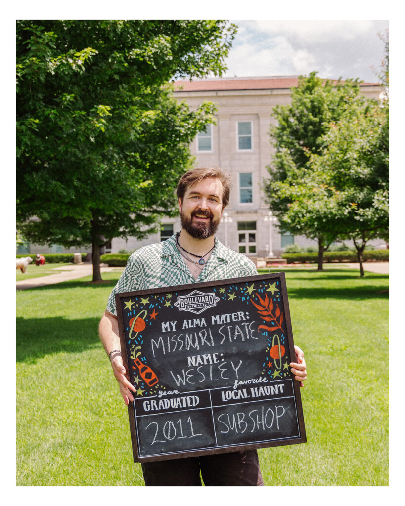 A smiling man stands on a grassy campus holding a chalkboard sign that reads: "My alma mater: Missouri State. Name: Wesley. Graduated: 2011. Favorite local haunt: Sub Shop." Trees and a building are in the background.