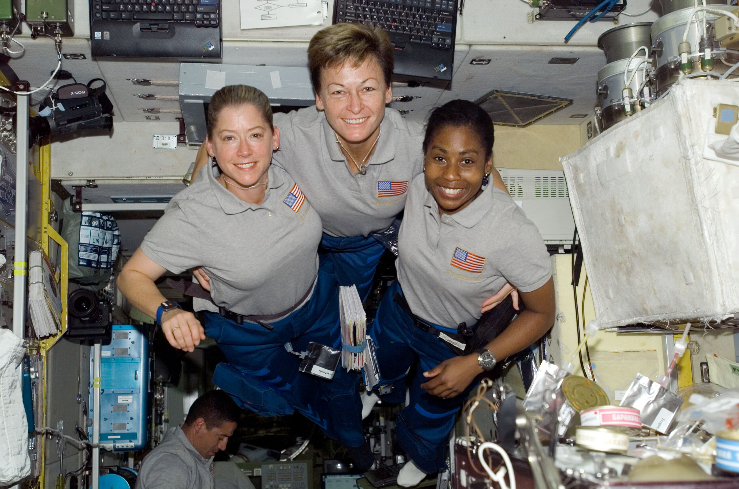 Three astronauts in gray shirts and blue pants float together inside a spacecraft, smiling at the camera. The interior is filled with equipment, cables, and supplies. Another crew member is partially visible below.