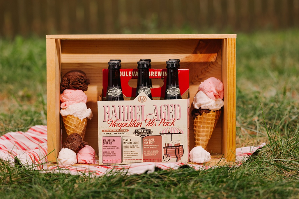 A wooden crate holds a pack of three bottled beers labeled “Barrel-Aged Neapolitan Mix Pack,” surrounded by three ice cream cones filled with chocolate, strawberry, and vanilla scoops, set on grass with a red-checkered cloth.