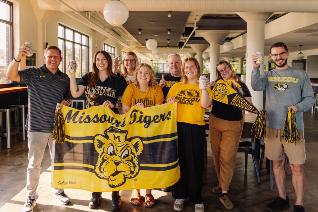 A group of eight people stand indoors holding drinks and Missouri Tigers gear, including a large yellow and black banner and pom-poms, all wearing University of Missouri apparel and smiling at the camera.