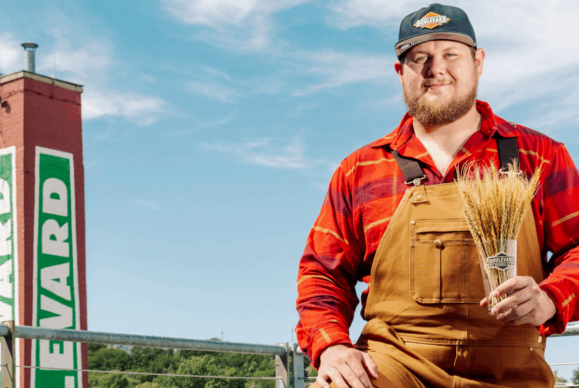 A man in brown overalls and a red plaid shirt holds a bundle of wheat. He stands outdoors in front of a tall red tower with "LEWARD" written vertically in green and white, under a blue sky.
