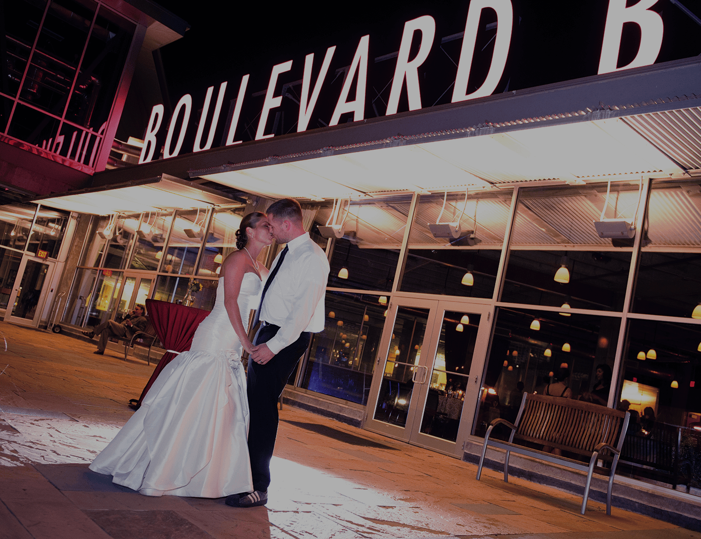 A bride and groom stand close together, holding hands and smiling at each other outside a brightly lit building with a large "BOULEVARD" sign at night.