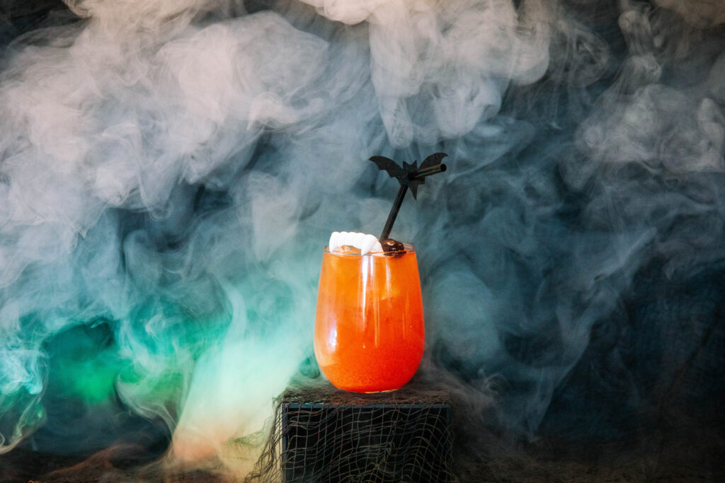 A bright orange Rec Deck the Halls cocktail in a glass with ice, garnished with a bat-shaped stirrer and white candy, sits on a dark surface surrounded by swirling fog and eerie lighting.