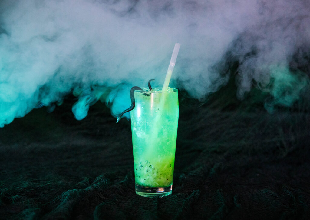A tall glass filled with a bright green, fizzy Rec Deck the Halls drink, garnished with a black gummy snake and a straw, sits on a dark textured surface with swirling fog in the background.
