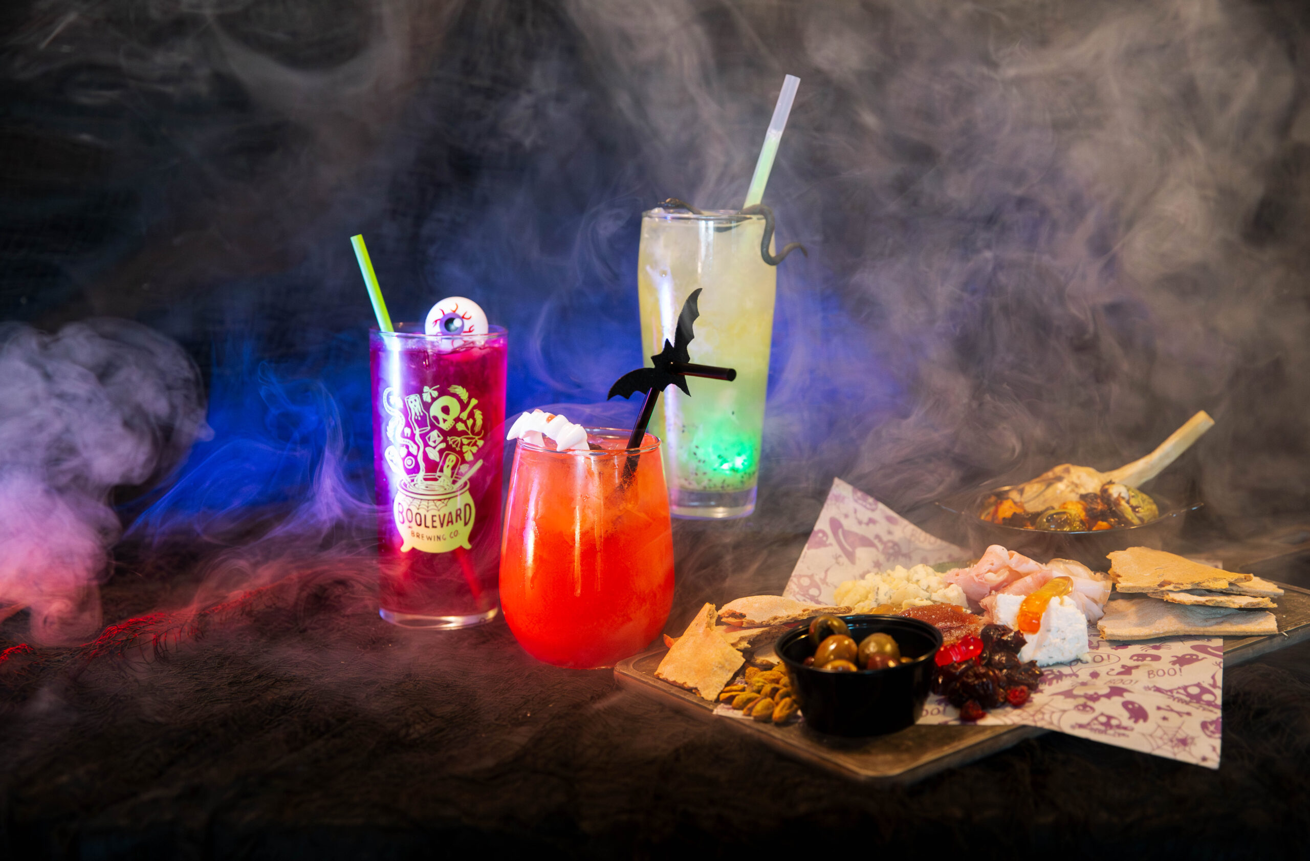 Three colorful cocktails with straws sit on a smoky, dark surface. In front is a platter with pita, cheese, olives, and snacks. Eerie blue and purple lighting creates a festive Halloween vibe.