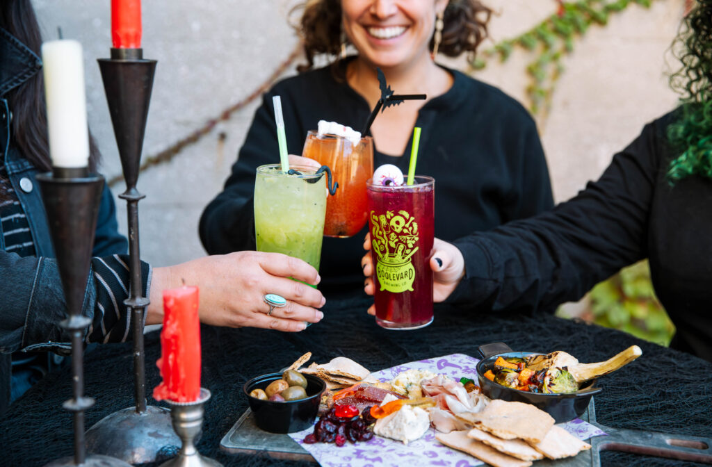 Three people clink colorful cocktails over a black table set with red candles and tasty snacks at a lively BOOlevard gathering. They are smiling and enjoying an outdoor celebration together.