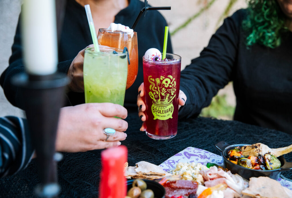 Three people clink colorful drinks—green, orange, and purple—over a table set with assorted food and Halloween-themed decor at Rec Deck the Halls. A fake eyeball garnish adds festive flair to one glass's creative design.