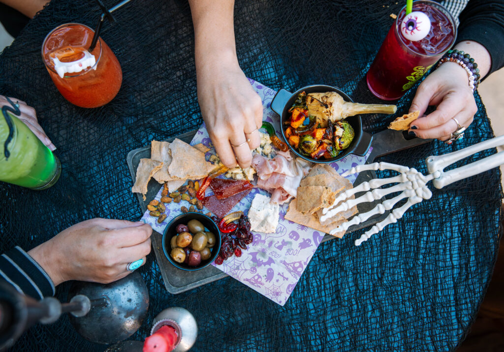 Three people’s hands (including one wearing a skeleton glove) reach for snacks like olives, crackers, and cheese on a table with drinks, creating a playful, spooky-themed Rec Deck the Halls meal setting.