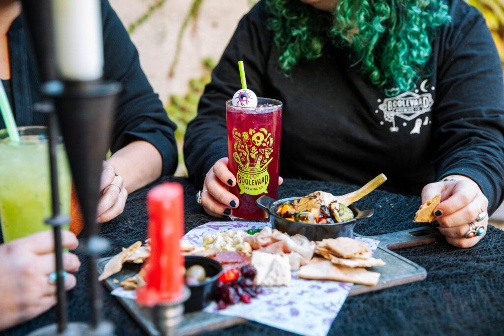 Two people at Rec Deck the Halls sit at a table with cocktails and a charcuterie board. One holds a purple drink in a Boulevard glass; the table is set with a candle, cheese, crackers, fruit, and other tempting snacks.
