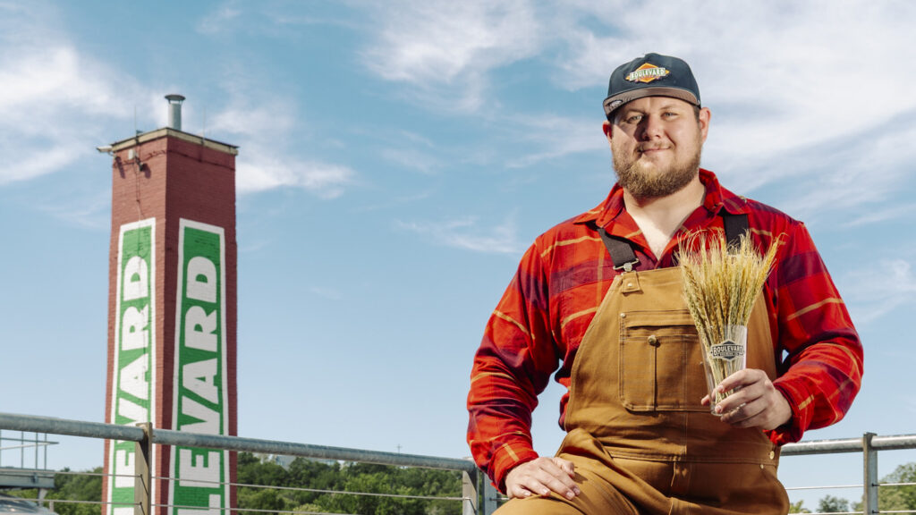 A man in brown overalls and a red plaid shirt holds a bundle of wheat. He stands outdoors near a red brick tower with “BLVD” painted on it, under a blue sky.