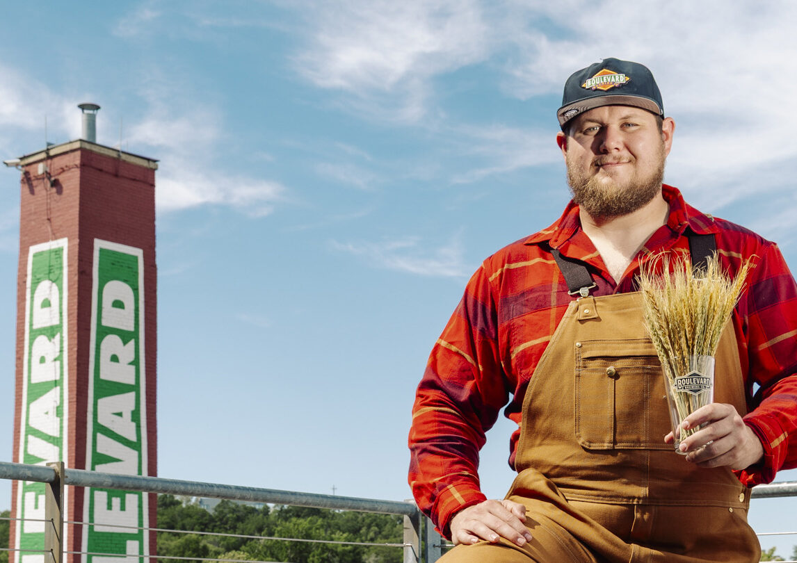 A man in brown overalls and a red plaid shirt holds a bundle of wheat. He stands outdoors near a red brick tower with “BLVD” painted on it, under a blue sky.