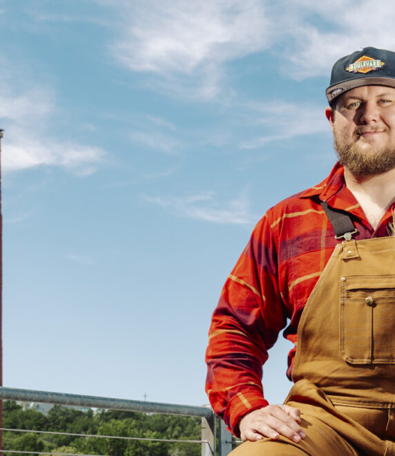 A man in brown overalls and a red plaid shirt holds a bundle of wheat. He stands outdoors near a red brick tower with “BLVD” painted on it, under a blue sky.