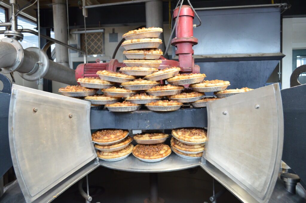 A large stack of freshly baked pies sits inside an open industrial baking oven in a factory setting, with machinery and metal pipes visible in the background.