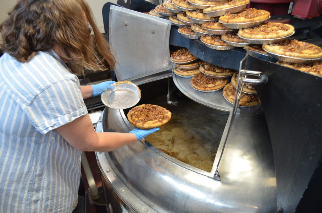 A person wearing blue gloves places a pie into a large, round industrial mash tun filled with pies and brewing beer, while many baked pies wait stacked on top of the vessel.