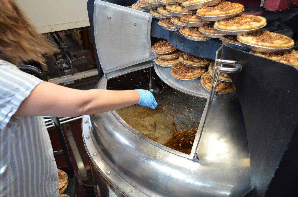 A person wearing a blue glove is operating a large, industrial machine that appears to be baking or processing multiple pies stacked on racks inside the equipment.