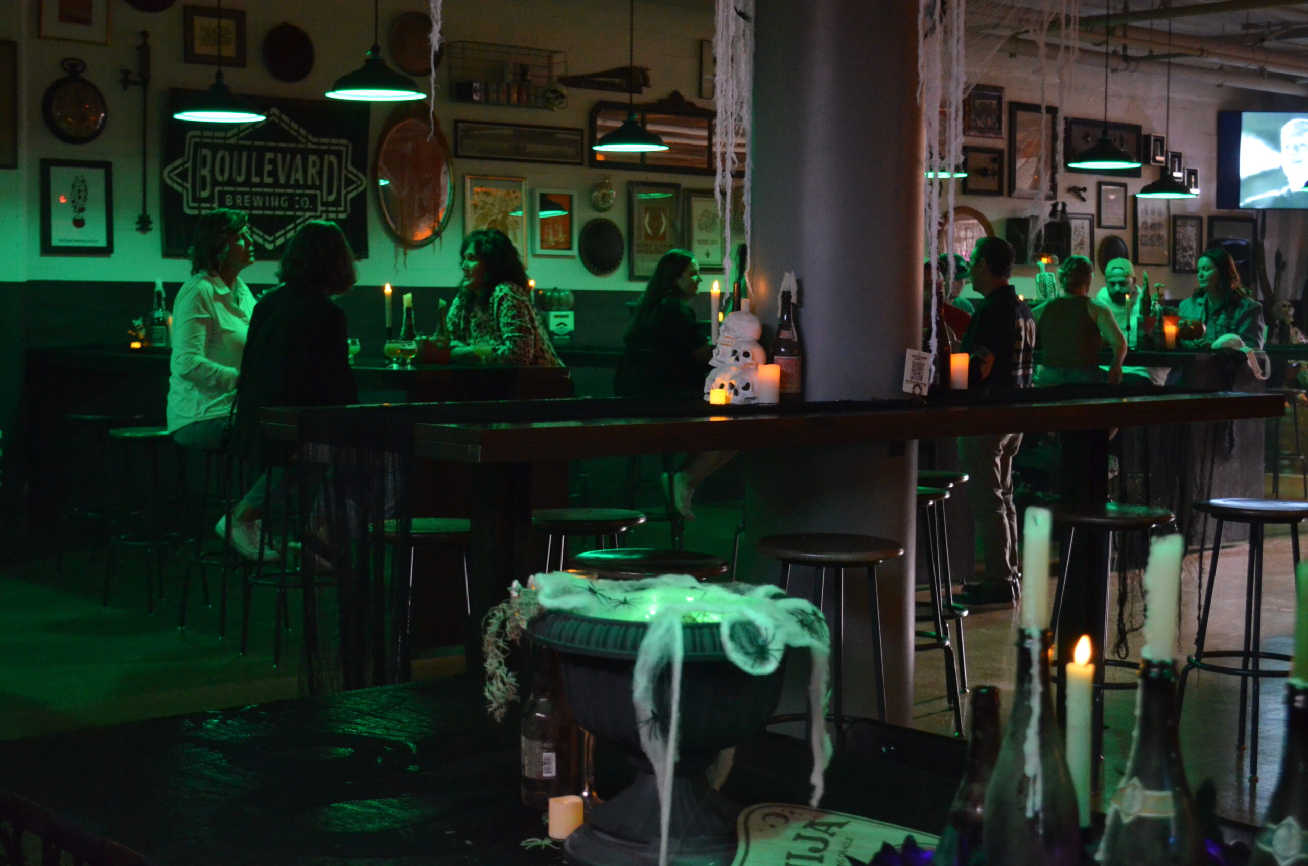 A dimly lit bar decorated for Halloween with cobwebs, candles, and pumpkins sets the scene for BOOlevard. People enjoy drinks under green-tinted lights while a Boulevard Brewing Co. sign glows on the wall.