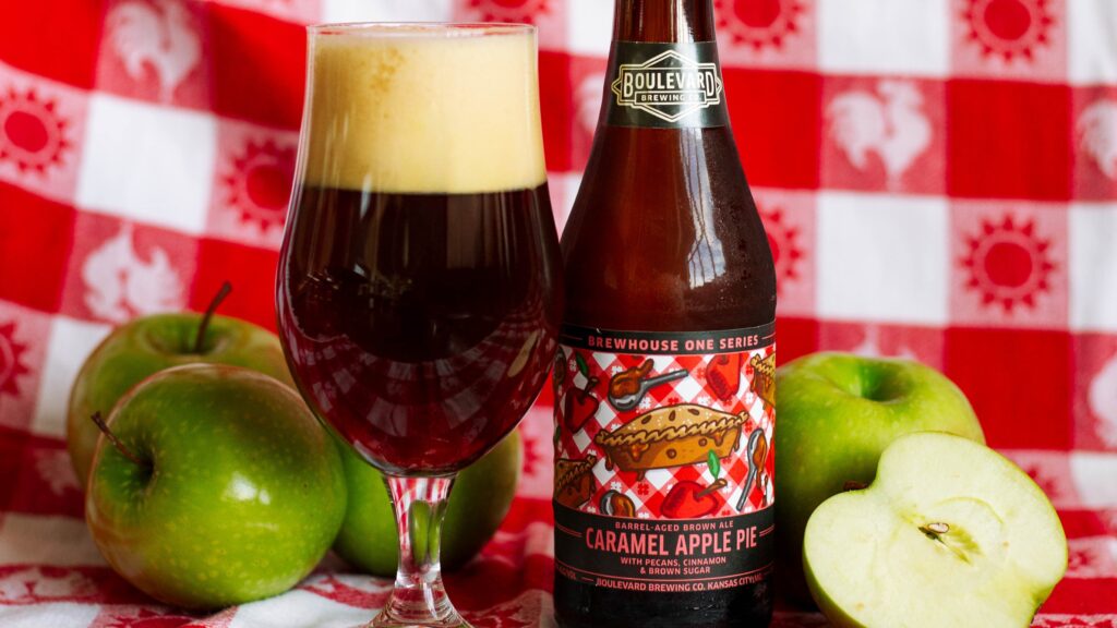 A bottle of Boulevard Brewing Co. Caramel Apple Pie beer and a glass of dark beer are placed on a red and white checkered cloth, surrounded by whole and sliced green apples.